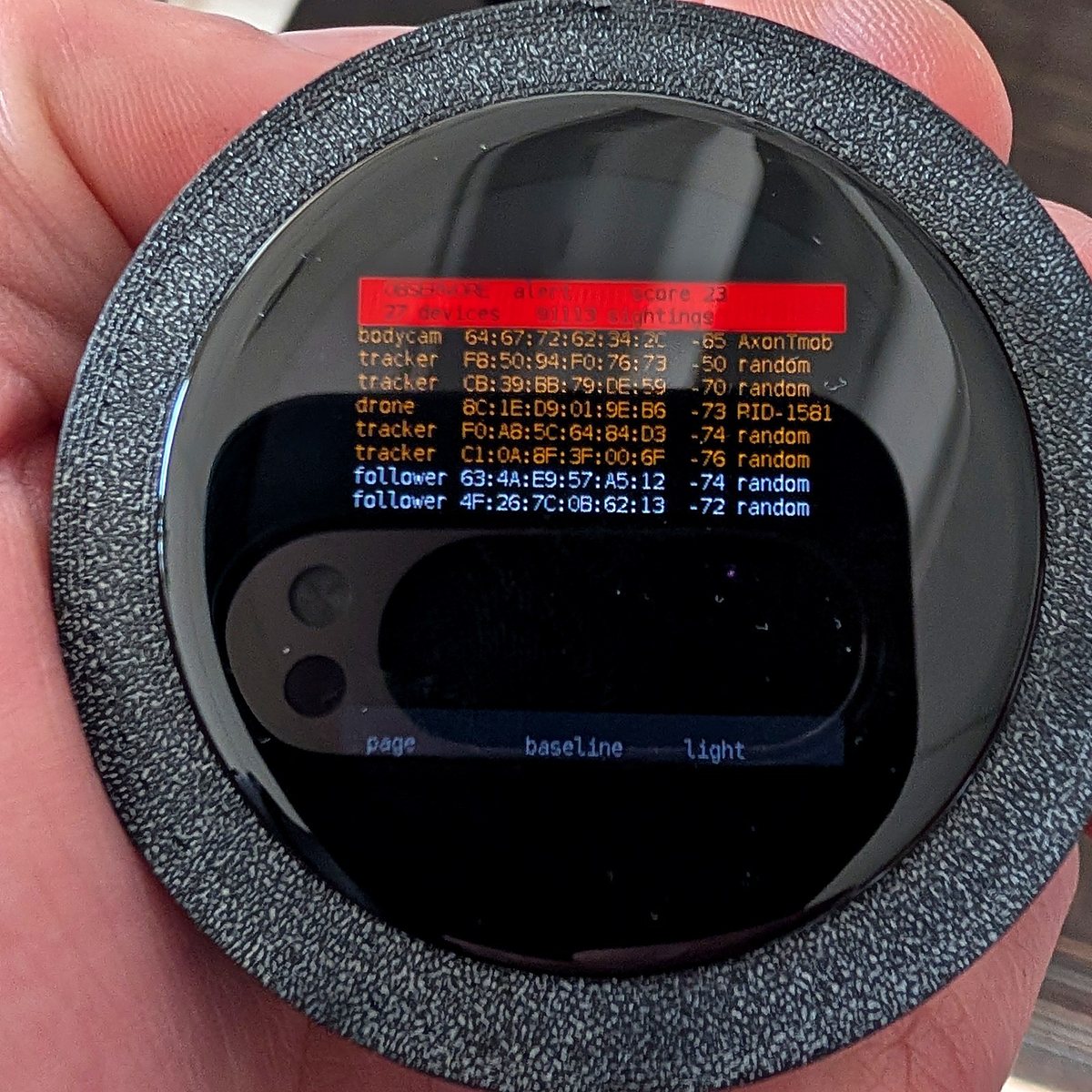 The round AMOLED board held in one hand, showing a red alert band reading score 23 above a list of findings: a body camera, a Remote ID drone, trackers and followers with signal strengths